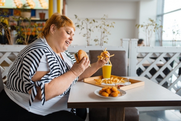 Fat woman eating high calorie food in fastfood restaurant. Overweight female person at the table with junk dinner, obesity problem