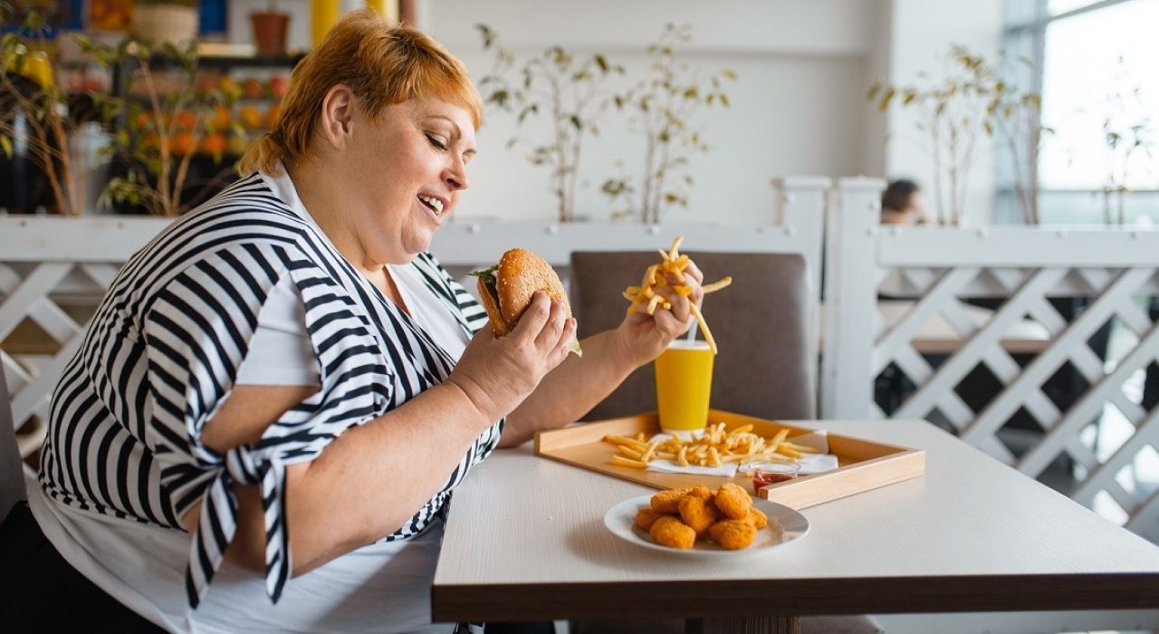 Fat woman eating high calorie food in fastfood restaurant. Overweight female person at the table with junk dinner, obesity problem