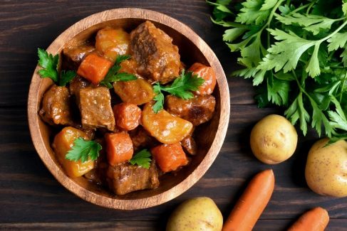 Meat stew with vegetables in bowl on wooden background, top view